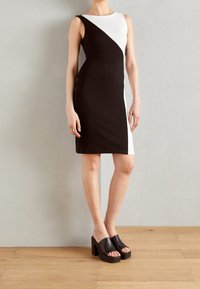 Kvinna i en ärmlös klänning i svart och vitt med färgblockering, i svarta plattformssandaler med öppen tå, stående på trägolv.