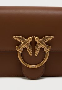 Bolsa de couro castanha com um fecho em tom de ouro e dois realces em forma de pássaros, além de detalhes costurados. Design minimalista com uma forma compacta.