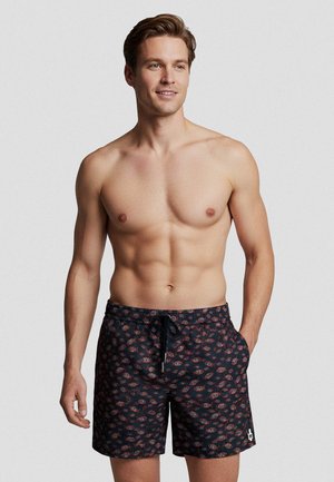 Lächelnder, oberkörperfreier Mann, der schwarze Badeshorts mit roten und blauen Blattmustern trägt, eine Hand in der Hosentasche, vor schlichtem hellem Hintergrund.