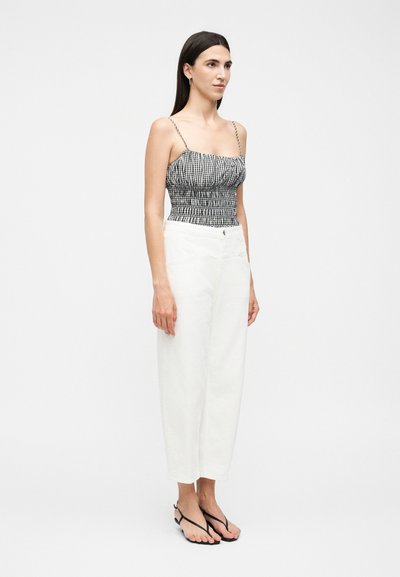 Γυναίκα με μακριά σκουρόχρωμα μαλλιά που φοράει ένα μαύρο και άσπρο καρό αμάνικο τοπ, λευκό ψηλόμεσο παντελόνι και μαύρα σανδάλια, στέκεται στραμμένη προς τα δεξιά.