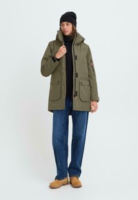 Olivově zelená parka s kapucí, na zip a na knoflíky, s několika předními kapsami, oblečená přes černý svetr, v kombinaci s modrými džínami a hnědou obuví.