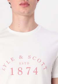 Tricou alb din bumbac, având un decolteu rotund și un design de text curbat de culoare roz care citește "LYLE & SCOTT ESTD 1874." Textură netedă.