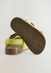 Gröna mockaskor med två justerbara remmar, texturerad brun gummisula och en korkliknande mellansula med vågig mönster.