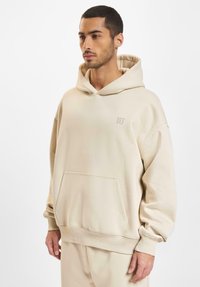 Beige hettegenser i myk bomullsblanding med kengurulomme, ribbestrikkede mansjetter og en subtil logo på brystet. Uformelt design og avslappet passform.