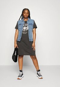 Denimväst över en svart t-shirtklänning med tryck; svarta sneakers med vita detaljer; svart axelväska; flätad frisyr. Mjuka texturer.