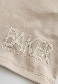 Beżowa bawełniana tkanina z dużym, fakturowanym logo „BAKER” z powtarzającym się wzorem „TED”, umieszczona blisko obrzuconego brzegu, z białymi akcentami szwów.