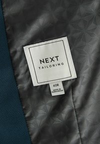 Tmavomodrá látka s textúrovaným povrchom, ktorá má šedé vzorované podšívkové vrstvu. Na bielej etike je napísané "NEXT TAILORING" a veľkosť "40R".