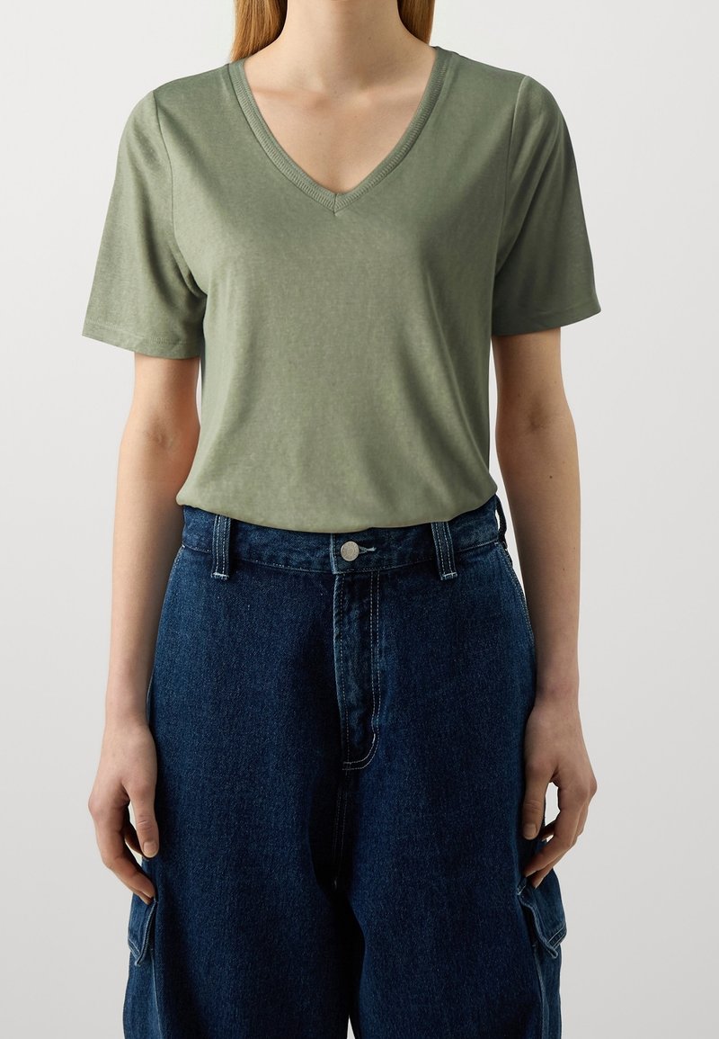 Personne portant un t-shirt uni vert clair à manches courtes avec col en V, rentré dans un pantalon en jean bleu taille haute avec poches latérales.