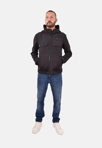 Zwarte zip-up jas met een capuchon, twee borstzakken en een soepele textuur. Gecombineerd met blauwe jeans en witte sneakers.