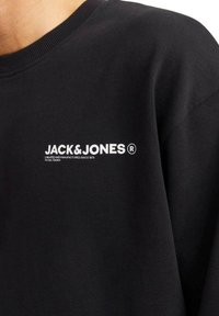 Sudadera negra con cuello redondo, puños acanalados y un estampado de logotipo blanco en el pecho, mostrando una textura de tela suave.