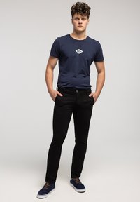 Granatowa koszulka z białym logo; czarne obcisłe dżinsy; niebieskie trampki z brązowymi sznurówkami; model stoi z rękami w kieszeniach na neutralnym tle.