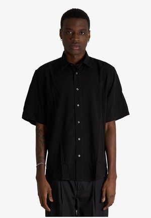 Giovane con capelli corti che indossa una camicia nera a maniche corte larga con bottoni e pantaloni neri, in piedi contro uno sfondo semplice.