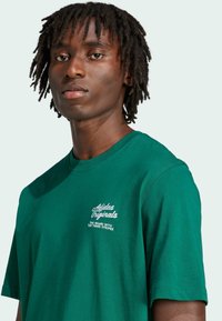 Green cotton t-shirt with short sleeves, featuring white embroidered text. Round neckline and relaxed fit, minimalistic design.