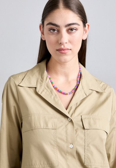 Jonge vrouw met lang donker haar, gekleed in een beige overhemd met knopen en een kleurrijke kralenketting, neutrale grijze achtergrond.