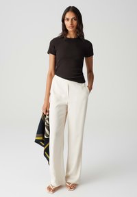 T-shirt nera aderente, pantaloni bianchi a vita alta e sandali beige. La modella tiene un foulard fantasia con dettagli blu navy, bianchi e dorati.