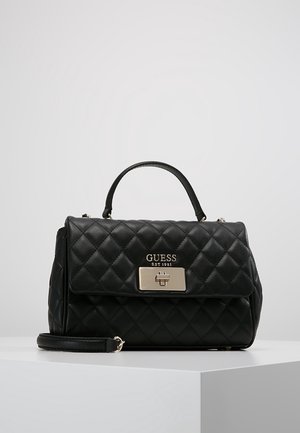 Black quilted leather handbag with top handle, gold-tone clasp, and GUESS logo on front flap, resting on a white surface.