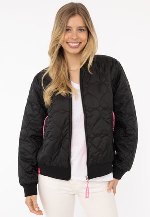 Lächelnde Frau, die eine schwarze Steppjacke mit Herzmustern, ein weißes Hemd und weiße Hosen trägt, vor einem schlichten Hintergrund stehend.