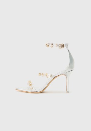 Sandalia de tacón transparente con tiras adornadas con tachuelas doradas, tacón delgado y diseño de punta abierta con un acabado suave y brillante.