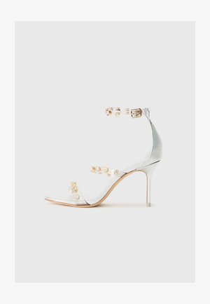 Sandalia de tacón transparente con tiras adornadas con tachuelas doradas, tacón delgado y diseño de punta abierta con un acabado suave y brillante.