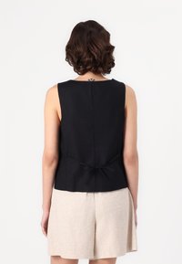 Top sem mangas preto com decote redondo e detalhe de laço nas costas. Apresenta uma textura suave e um corte ajustado. Combinado com calções bejes claros.