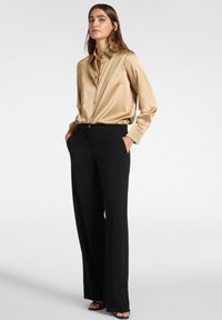 Eine Frau steht mit einem goldenen Satin-Knopfhemd, das in eine hochtaillierte schwarze weit geschnittene Hose und schwarze Absatzsandalen gesteckt ist.