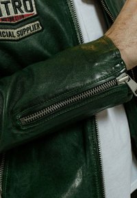 Veste en cuir vert avec fermeture éclair en métal, surface texturée et écusson portant la mention "MOTRO". Elle se caractérise par une coupe ajustée et une finition douce.