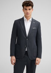 Abito grigio scuro su misura con finitura texturizzata, dotato di due tasche e chiusura con un bottone. Indossato sopra una camicia bianca.