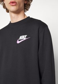 Jeune personne aux cheveux bouclés portant un sweat-shirt noir Nike avec un logo swoosh rose sur la poitrine, sur un fond uni.