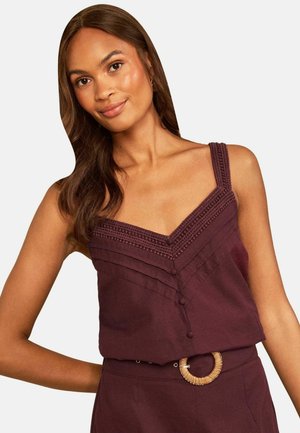 Vrouw met lang bruin haar die een mouwloze donkerpaarse top en bijpassende broek draagt met een geweven ronde riemgesp.