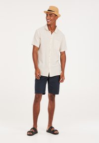 Korte mouwen, beige gestreept shirt met een zak, gecombineerd met marineblauwe shorts. Het model draagt zwarte sandalen en een strohoed met een zwarte band.