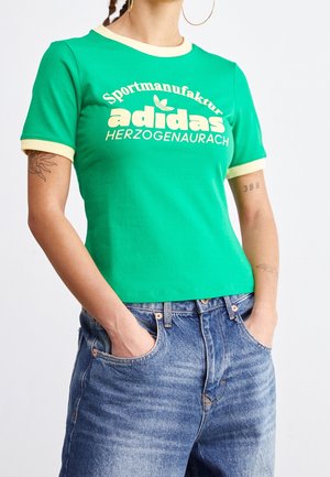 Person iført grøn Adidas t-shirt med teksten "Sportmanufaktur HERZOGENAURACH", blå jeans og øreringe i ringform, hænderne i lommerne.