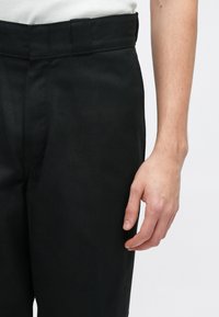 Svarte bomullsbukser med en midjebånd vises, og viser en jevn tekstur og en tettsittende silhuett, sammen med en hånd som hviler på stoffet.