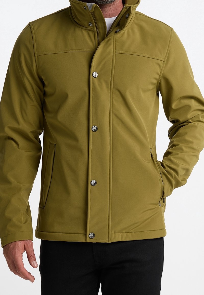 Man i olivgrön jacka med knappar framtill och sidofickor med dragkedja, i kombination med svarta byxor, händer delvis synliga.