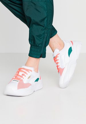 Persona che indossa sneaker bianche Puma con accenti rosa e verdi e lacci corallo, abbinate a pantaloni jogger verde scuro con cerniere alla caviglia.
