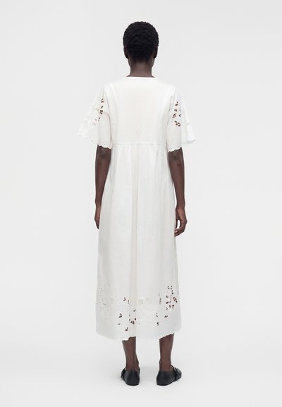 Personne portant une robe midi blanche à manches courtes avec broderie florale découpée, debout de dos sur un fond uni.