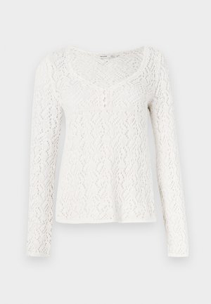 Chemise blanche en dentelle à manches longues avec un col rond, présentant un motif en losanges texturé et de petits boutons sur le devant.