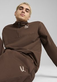 Brun zip-up jacka med en texturerad yta; har en logotyp på bröstet och matchande bruna byxor med logotyp på låret.