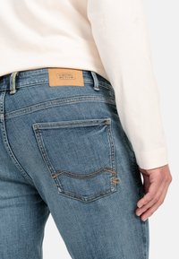 Blå denimjeans med et struktureret stof, der har et lædermærke, gule syninger og to baglommer med buede sømme.
