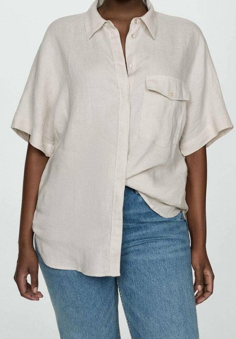 Personne portant une chemise beige ample à manches courtes avec une poche avant et un jean bleu, sur un fond uni.