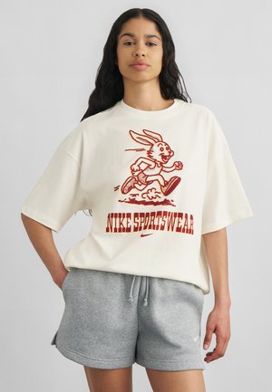Γυναίκα με μακριά σκούρα μαλλιά φοράει υπερμεγέθη λευκό T-shirt με τρέχον κουνέλι και το κείμενο "NIKE SPORTSWEAR", συνδυασμένο με γκρι σορτς.