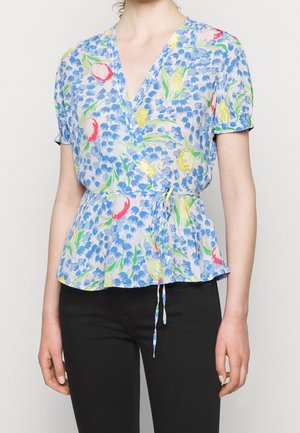 Vrouw draagt een kortemouws wikkelblouse met blauwe, gele en rode bloemenprint, gestrikt in de taille, gecombineerd met een zwarte broek.