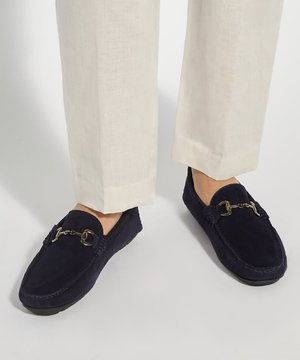Mocassins en suède bleu marine avec détail en métal, portés avec un pantalon large beige sur un fond uni.
