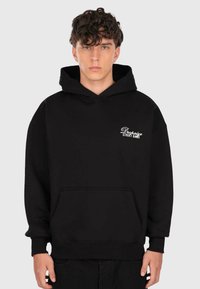 Jeune homme aux cheveux bouclés portant un sweat à capuche noir avec le texte blanc "Dropsize Street Game" sur la poitrine gauche, se tenant devant un fond uni.