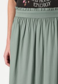 Gonna verde chiaro a vita alta con tessuto arricciato e cinturino elastico. Abbinaa a una maglietta nera con grafica che presenta testo e design.