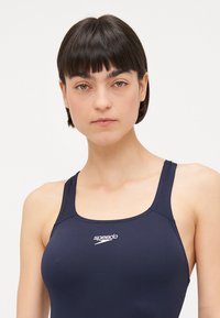 Maillot de bain bleu marine avec un design à dos nageur, fabriqué en matière lisse. Affiche le logo "Speedo" en blanc sur la zone de la poitrine.