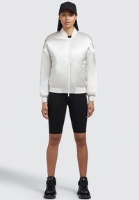 Giacca bomber in satin argento con polsini e orlo a costine, abbinata a pantaloni bike neri e sneakers alte nere. Cappellino nero indossato all'indietro.