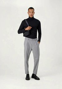 Black turtleneck, light gray pleated trousers, and black shoes. Model holds a small black bag, standing against a neutral background.