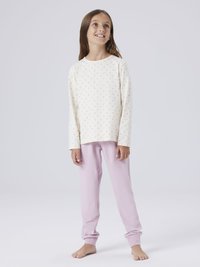 Jeune fille portant un haut à manches longues à fleurs claires et un pantalon rose, debout pieds nus sur un fond blanc uni, regardant vers le haut et souriant.