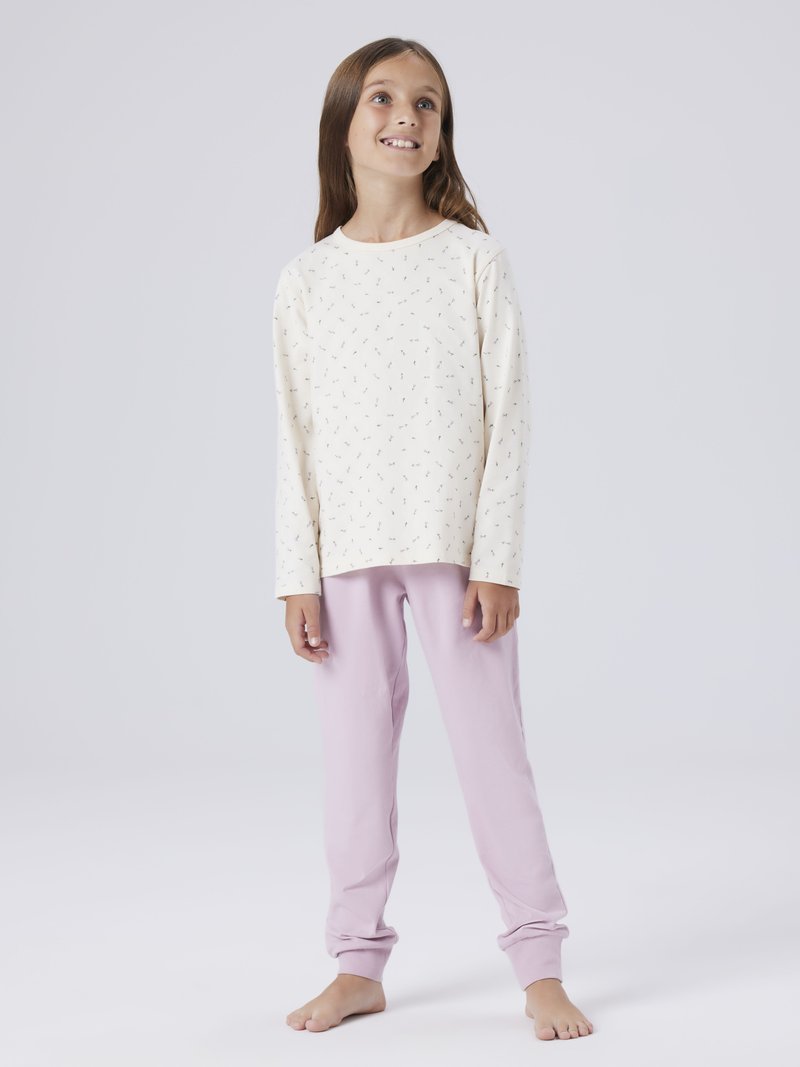 Jeune fille portant un haut à manches longues à fleurs claires et un pantalon rose, debout pieds nus sur un fond blanc uni, regardant vers le haut et souriant.