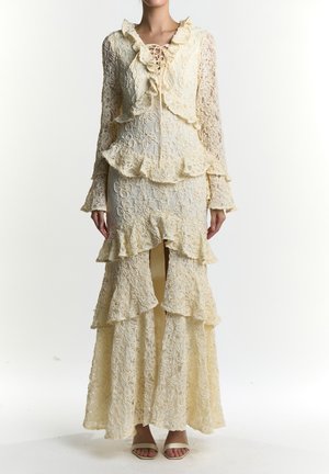 Koronkowa sukienka w blady żółty kolorze z falbankami, długimi rękawami i wiązaniem z przodu. Charakteryzuje się warstwową konstrukcją i wysokim rozcięciem przy dole.